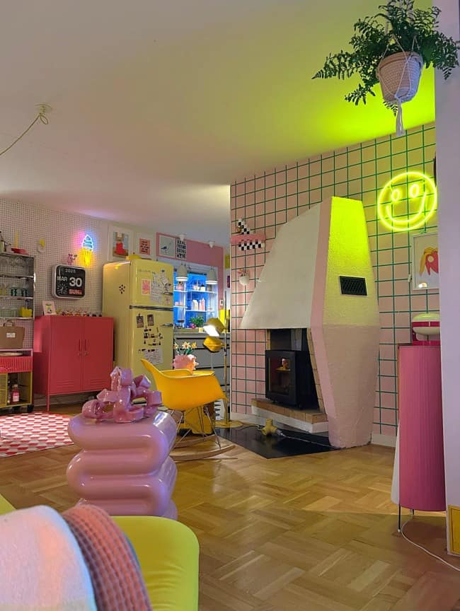 A retro, pastel-infused open concept living room displaying maximalist interior design, featuring a blocky pink and white stone fireplace setup against a pink-and-green gridded accent wall, complete with a neon smiley face sign and a vintage yellow refrigerator.