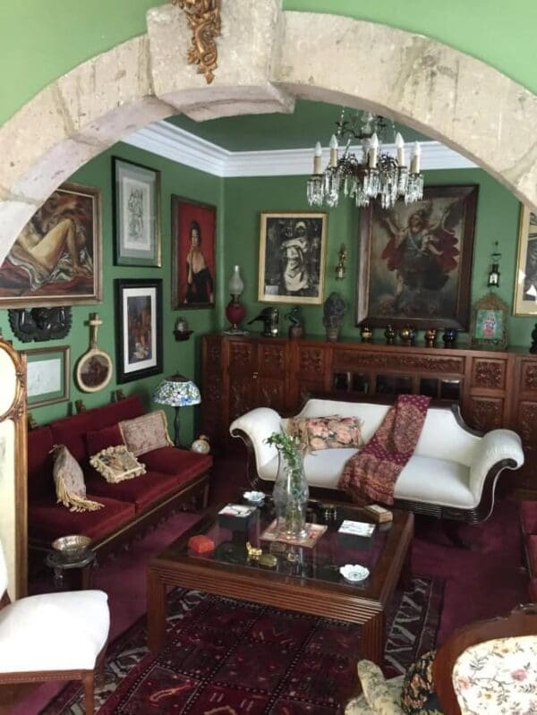 A classic, dark-toned parlor room framed by a stone archway illustrating historic maximalist interior design, with deep forest green walls crowded by ornate gold-framed classical oil paintings, a rich crimson velvet sofa, and a crystal chandelier.