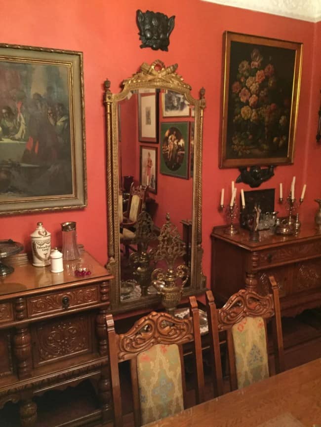 A moody dining room corner showcasing antique maximalist interior design, where deep terracotta red walls are adorned with classical portraits and a large gilded gold mirror that reflects dark, carved wood sideboards topped with vintage brass candlesticks.