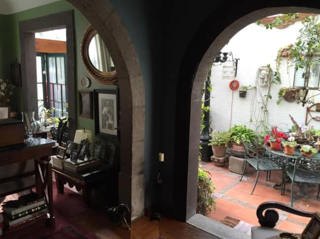 A view looking through heavy, rustic stone archways from a dark green interior sitting room filled with vintage frames out onto a sun-bleached white stone patio courtyard overflowing with potted plants and bright red flowers.