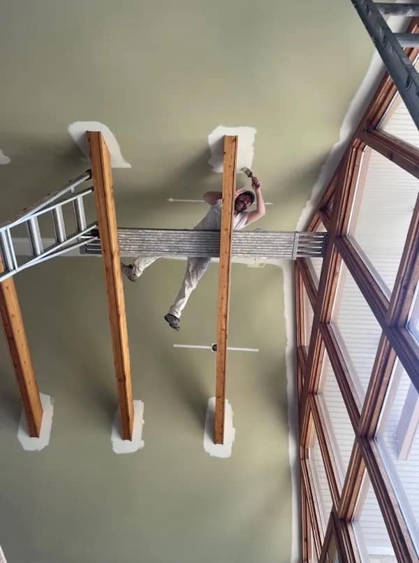 An absolute disaster of workplace safety in this funny meme showing a house painter precariously balancing an aluminum scaffold plank directly between two exposed ceiling beams while standing right over an open drop.