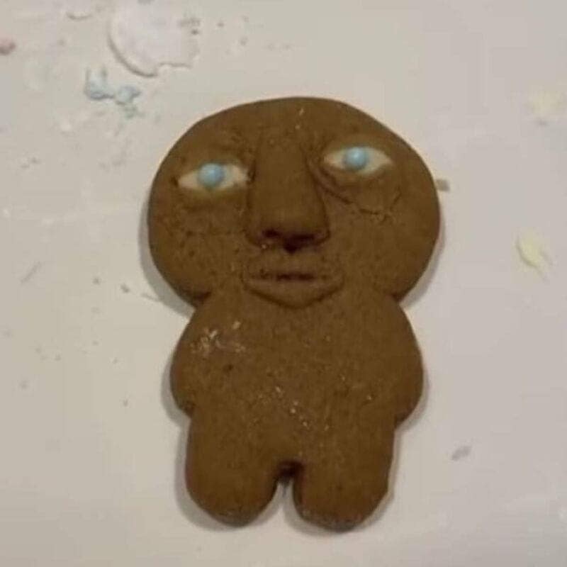 A cursed image from a chaotic meme dump showcasing a gingerbread man cookie baked with a disturbingly realistic human face, complete with a prominent nose, defined lips, and wide, unblinking light-blue eyes.