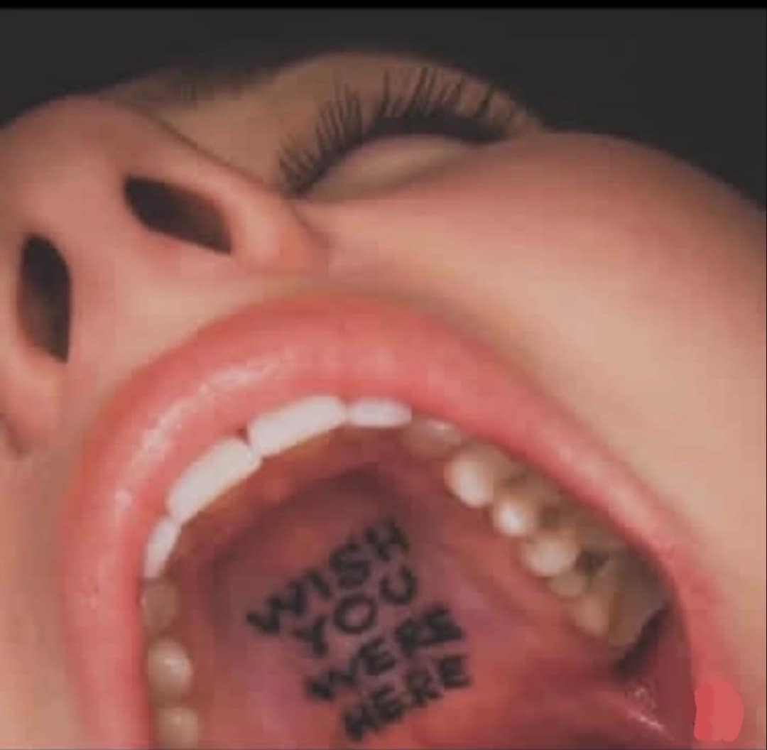 : A surreal close-up shot of an open human mouth from a shocking funny meme, revealing the phrase "WISH YOU WERE HERE" tattooed directly onto the back of the throat and tongue.