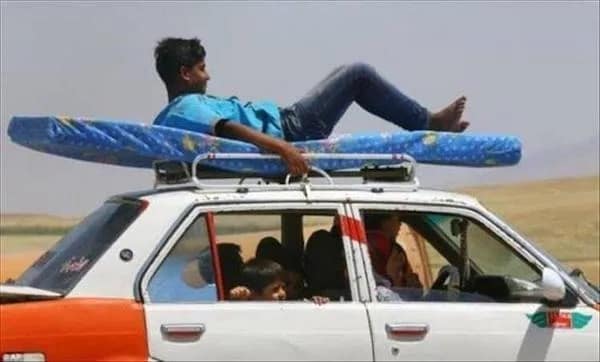 Boy riding on a blue mattress strapped to the roof rack of a white and orange moving sedan in a funny transit meme dump photo.