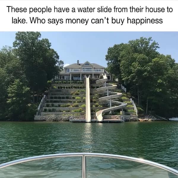 A stunning luxury real estate meme from a meme dump showing a massive multi-million dollar mansion built on a steep, perfectly manicured terraced hillside overlooking a lake. Two massive, resort-style white water slides—one a terrifying straight drop and the other a massive spiral loop—lead directly from the house into the water.
