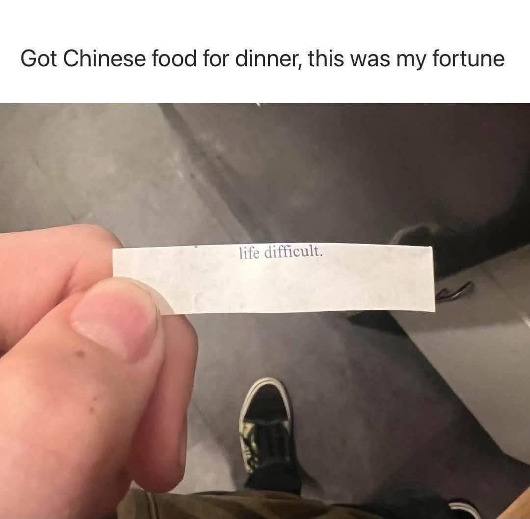 A close-up photograph from a relatable meme dump showing a hand holding a tiny white slip of paper from a fortune cookie that completely skips any future prediction to simply state: "life difficult."