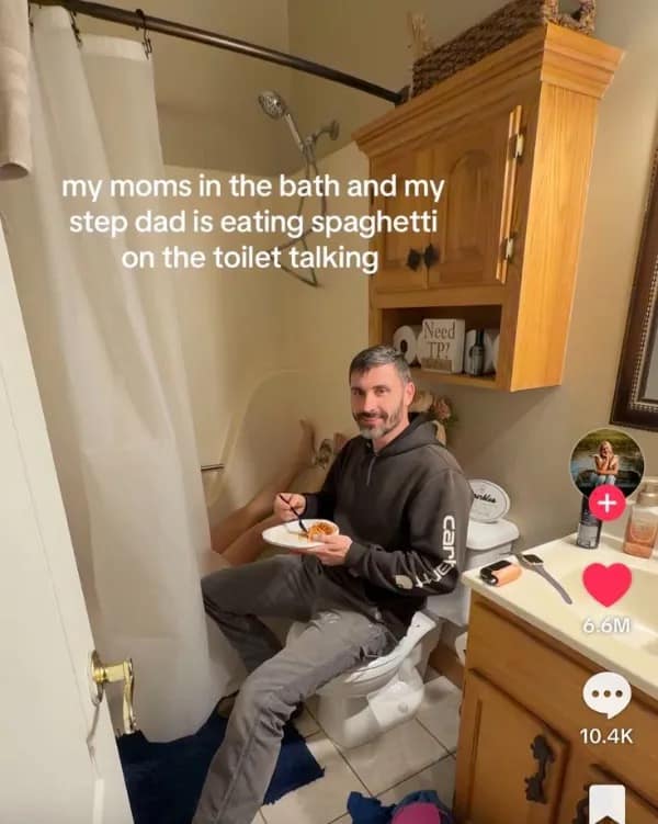A chaotic domestic meme from a meme dump displaying a viral TikTok screenshot inside a small bathroom. A casual stepdad in a gray hoodie sits directly on the closed toilet bowl calmly eating a plate of spaghetti while chatting with the mother, whose bare feet are poking out of the adjacent bathtub behind a plastic shower curtain.