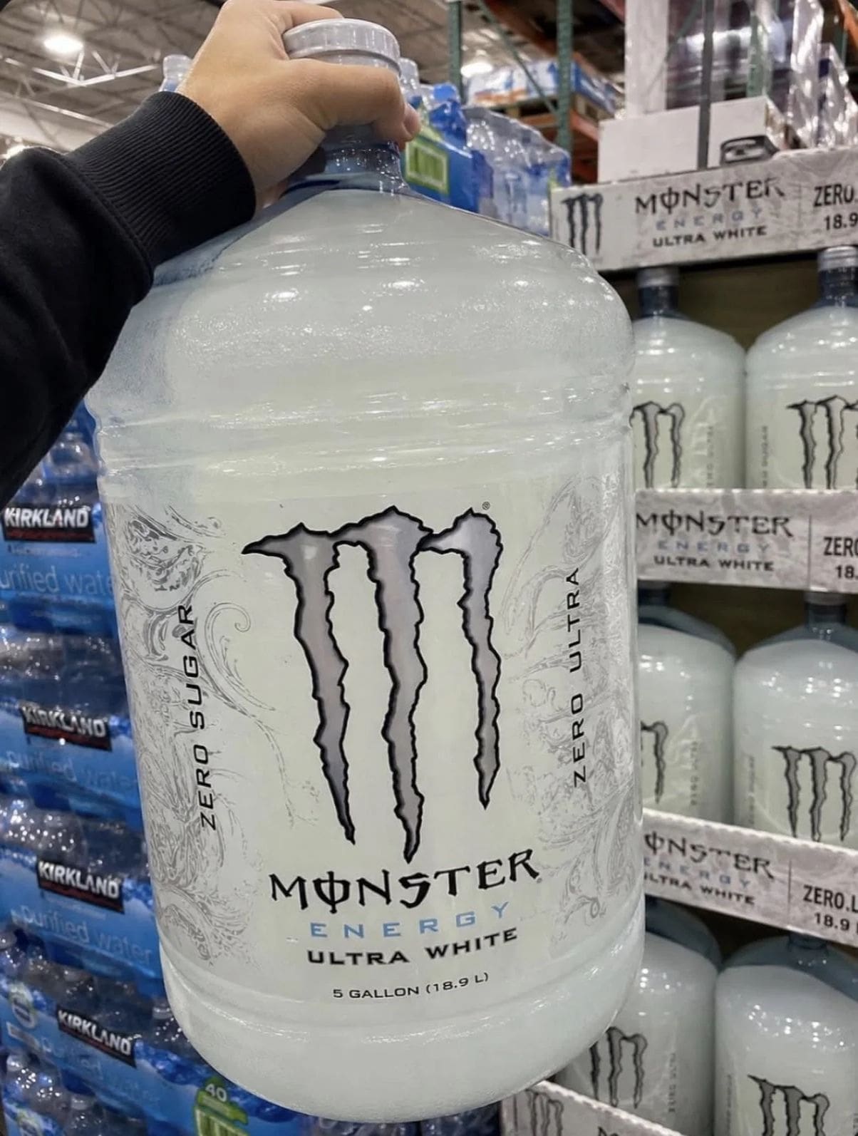 An item from a gallery of funny memes depicting a giant 5-gallon water-cooler style jug branded identically to a Monster Energy Ultra White can, held up by a shopper in a wholesale grocery aisle.