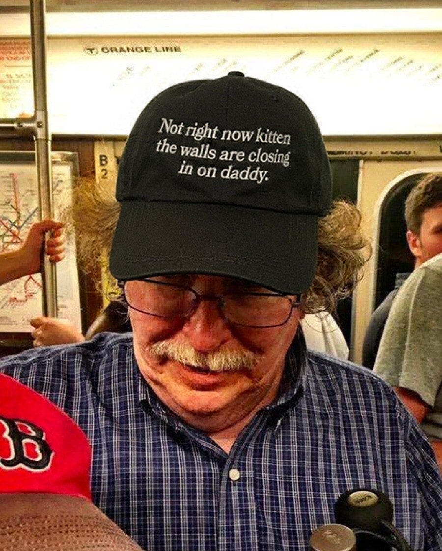 A public transit photograph common in chaotic memes, showcasing a middle-aged man with a mustache on a crowded subway train wearing a black dad hat embroidered with the unhinged phrase: "Not right now kitten the walls are closing in on daddy."