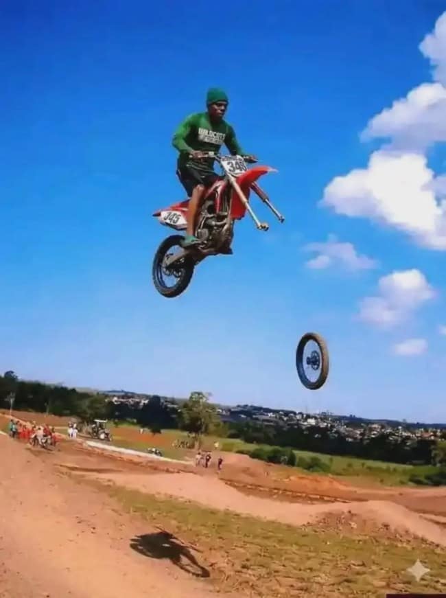 A dramatic instance of moments before disaster, capturing a motocross dirt bike rider suspended high mid-air against a bright blue sky as his front wheel completely detaches and plummets below his fork tubes.