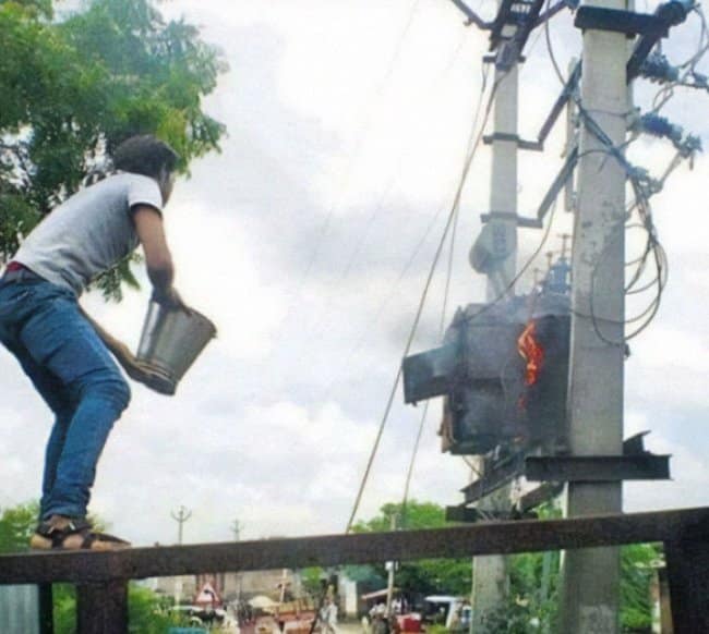 A wildly dangerous moments before disaster scene showing a man standing on an elevated ledge, rearing back to throw a metal bucket of water directly onto a massive electrical transformer box that is actively shooting sparks and catching fire.