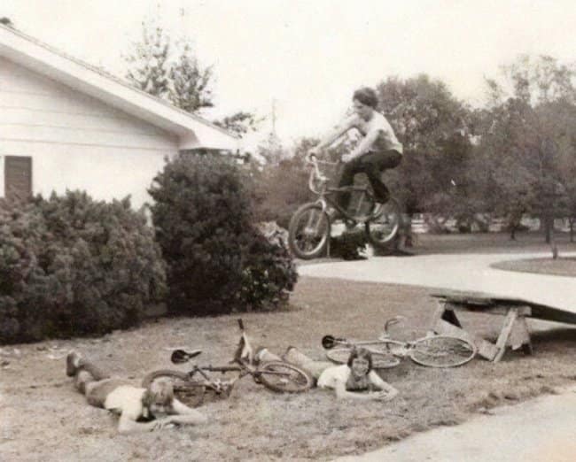 A vintage black-and-white moments before disaster photo showing two kids lying flat on the grass while a teenager jumps a BMX bicycle off a wooden ramp, currently suspended mid-air directly over their unhelmeted heads.