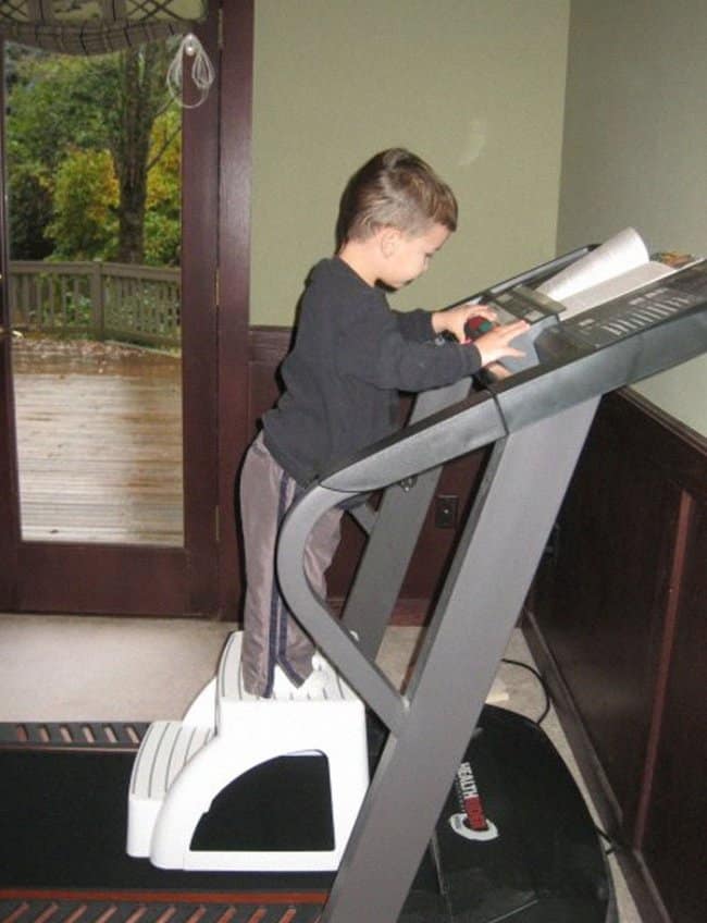 A child-safety moments before disaster frame capturing a toddler standing barefoot on a small plastic step stool placed directly on the motorized belt of an active treadmill, reaching up to touch the console buttons.