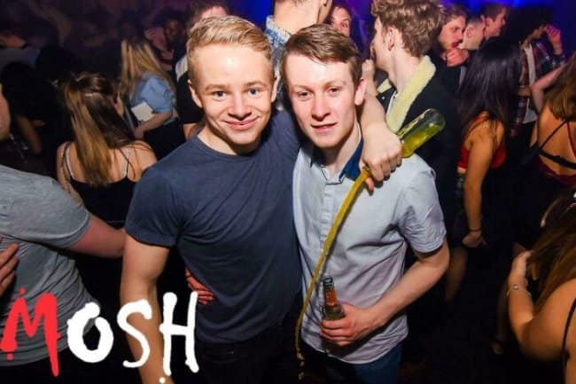 A chaotic nightclub moments before disaster photo showing two young men posing for a picture, completely unaware that a woman behind them is tilting a full glass bottle, pouring a thick stream of yellow liquid directly down the front of the man's shirt.
