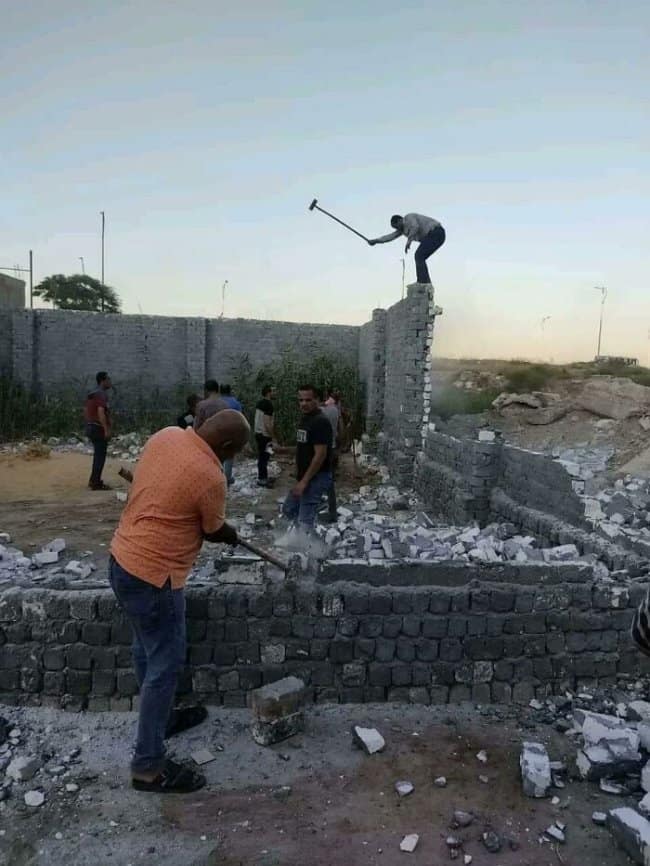 A terrifying construction moments before disaster image showing a man standing at the very top edge of a tall, unsupported brick wall, swinging a heavy sledgehammer down onto the exact brick he is currently standing on.