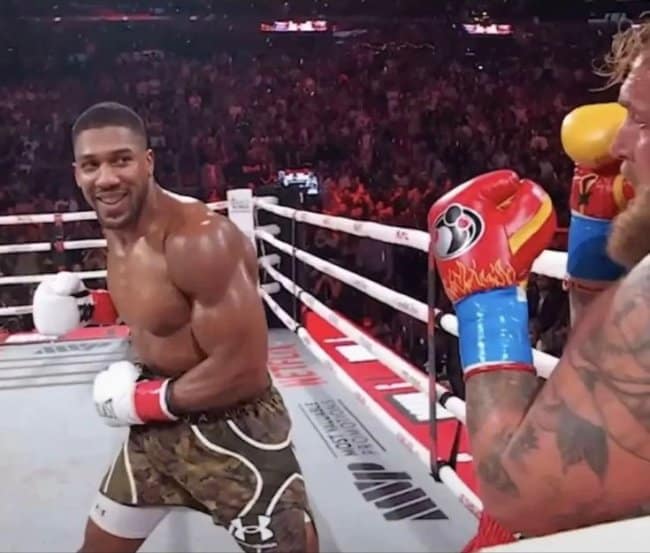 A sporting moments before disaster snapshot inside a boxing ring, showing fighter Anthony Joshua smiling warmly away from his opponent while a massive red glove with flame patterns sails directly toward his face in the immediate foreground.