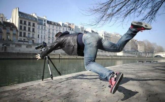 A frantic photography moments before disaster shot capturing a man lunging forward horizontally along a stone riverbank, desperately trying to catch his expensive camera and tripod before it tips entirely into the water below.