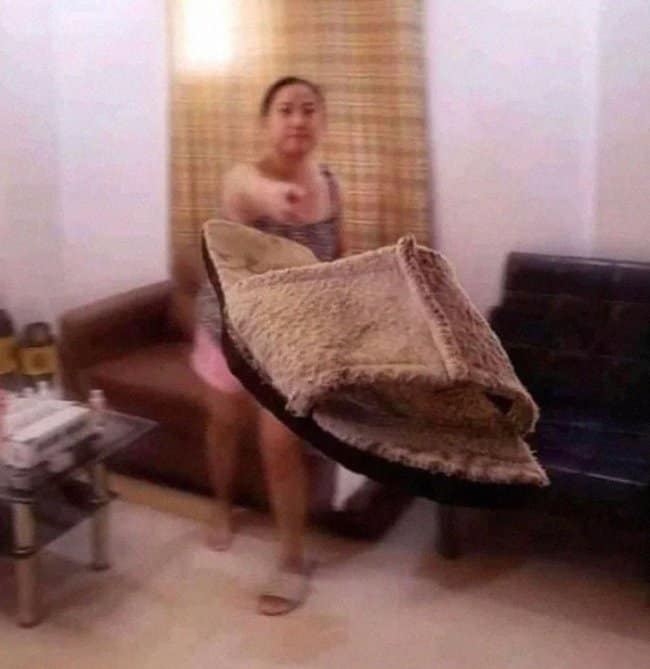 A chaotic, motion-blurred moments before disaster frame capturing a woman inside a living room violently launching a large brown plush pet bed directly toward the camera lens.