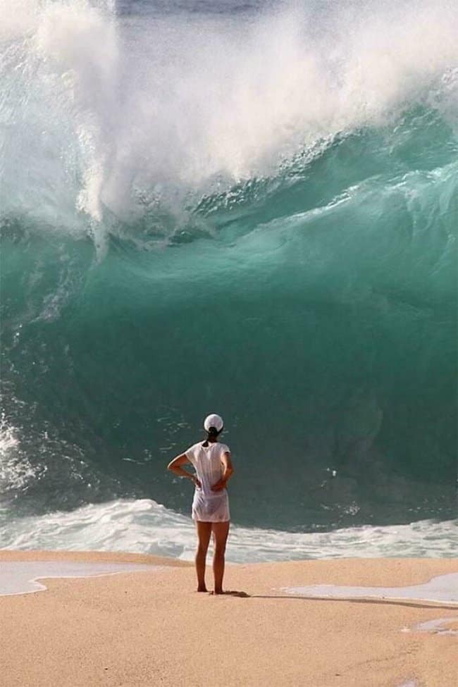 A breathtakingly intense shoreline moments before disaster scene showing a woman standing with her hands on her hips, casually staring up at a monolithic, towering turquoise shorebreak wave about to crash over her.