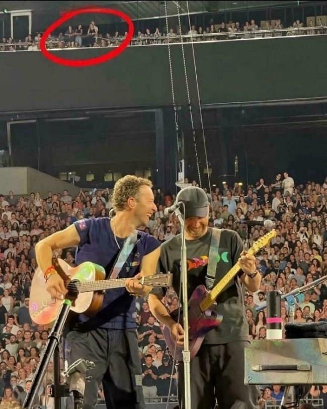 A stadium concert view operating as a moments before disaster viral meme template, showing Chris Martin of Coldplay performing on stage while a red circle highlights a reckless spectator falling or hanging dangerously over the upper balcony railing.
