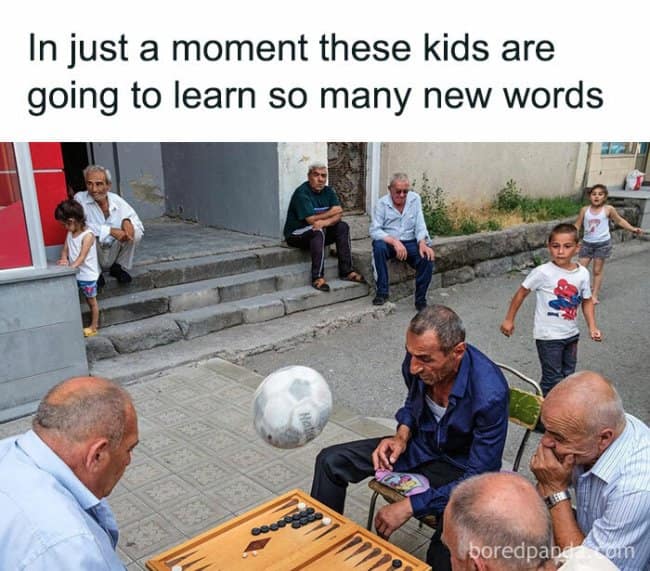 A text-captioned street photo framing a literal moments before disaster, where a stray soccer ball is frozen mid-flight inches away from hitting an elderly man playing backgammon, topped by the text: "In just a moment these kids are going to learn so many new words".