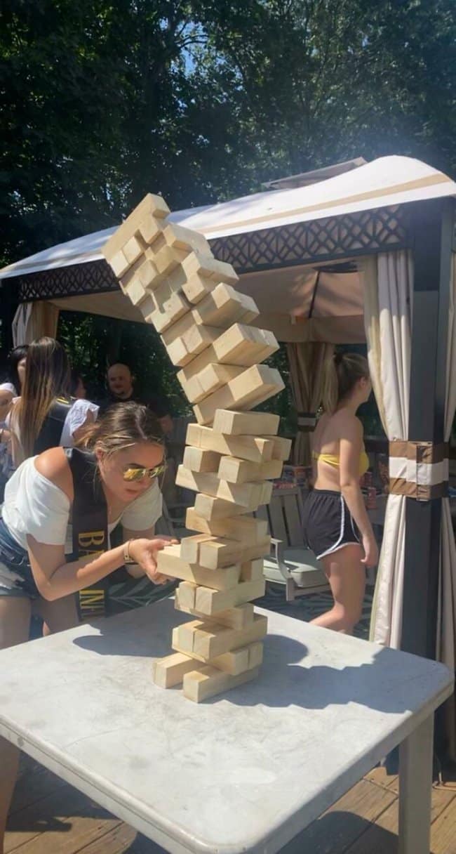 A tense backyard party snapshot framing moments before disaster, capturing a woman trying to delicately extract a block from a giant wooden Jenga tower that is actively bowing, twisting, and mid-collapse.