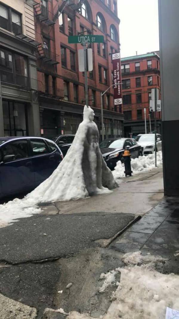 A massive snow sculpture shaped like Batman standing on a city sidewalk underneath a street sign.