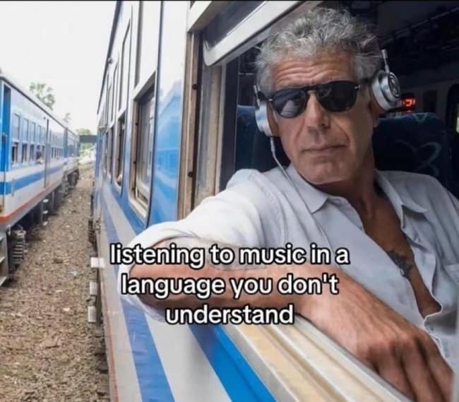 A travel-themed music meme featuring Anthony Bourdain wearing headphones and sunglasses while looking out of a train window, captioned, "listening to music in a language you don't understand."