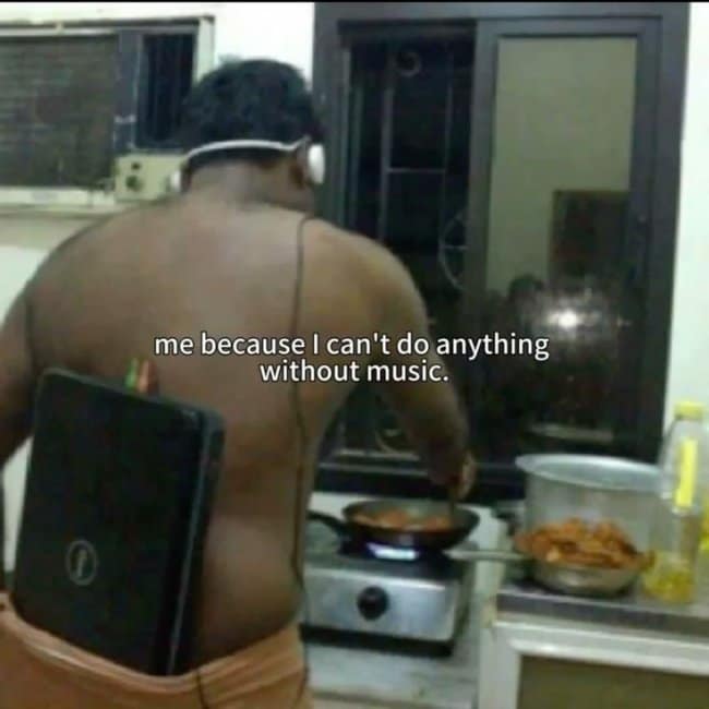 An absolute dependency music meme showing a shirtless man in a kitchen cooking over a stovetop with a massive black laptop tucked directly into the back of his waistband to keep his headphones plugged in.