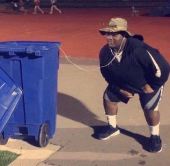 A funny listening habits music meme capturing a person in a sun hat bending over on an outdoor track to listen closely to a cord running directly inside an open blue plastic trash bin.