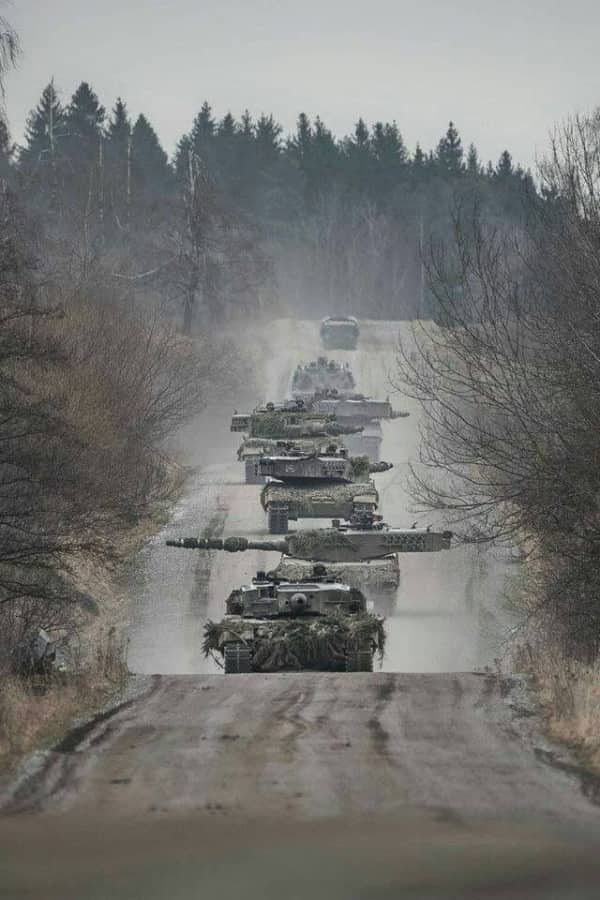 Line of heavily camouflaged military tanks driving down a dirt road through a barren forest.