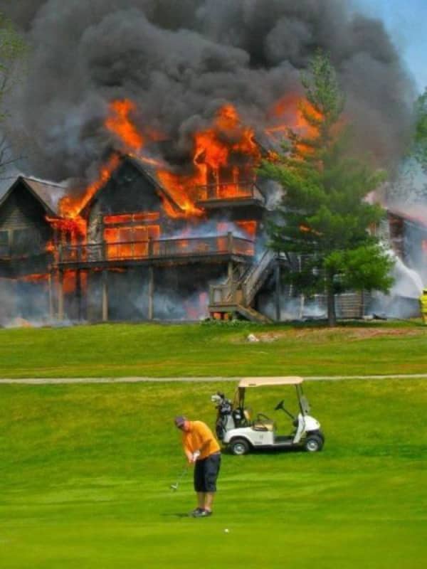 Man focused on putting a golf ball on a green while a large building burns in background.