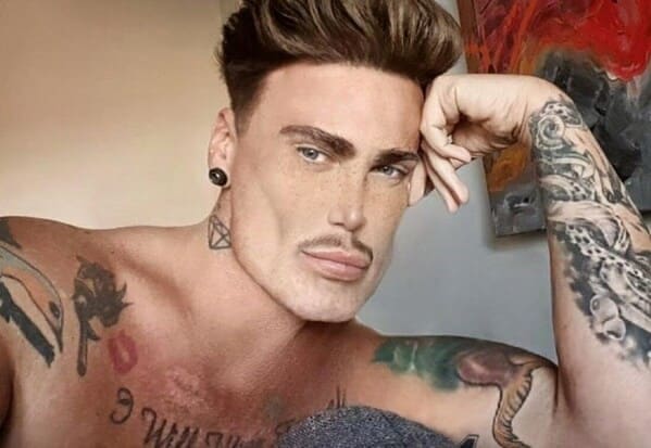 A close-up selfie of a tattooed man with ear gauges, displaying an extreme facial photoshop fail where his jawline and cheekbones have been comically morphed into a razor-sharp, cartoonish "GigaChad" aesthetic.