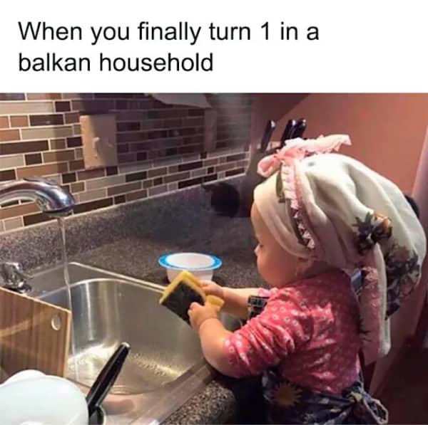 A toddler wearing a traditional floral headscarf wash dishes by hand inside a kitchen sink.