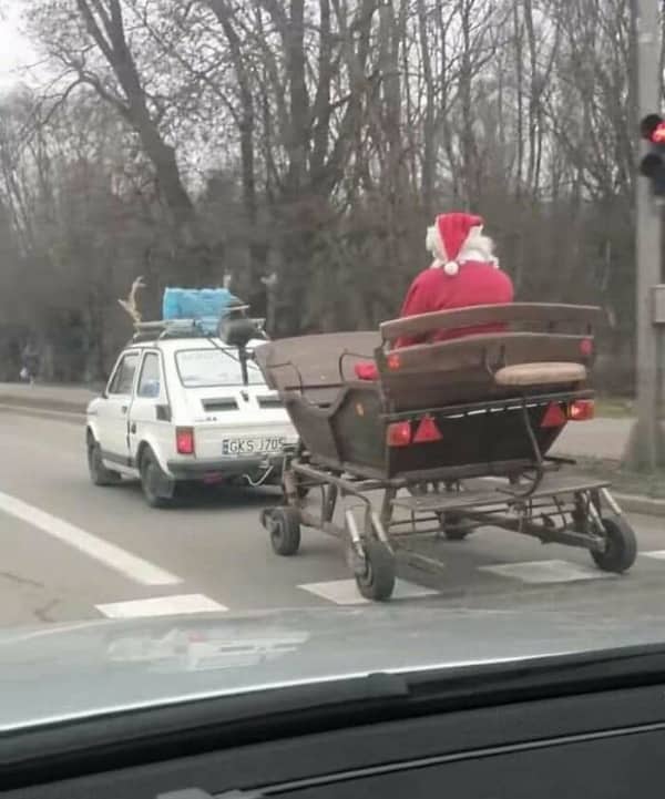 A person dressed as Santa Claus riding inside a wooden carriage towed behind a tiny white car.