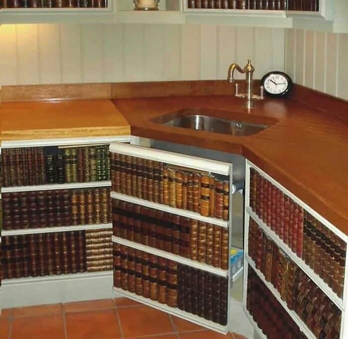 A corner kitchen sink counter with cabinets underneath disguised as rows of antique library books, highlighting a unique instance of weird home decor.