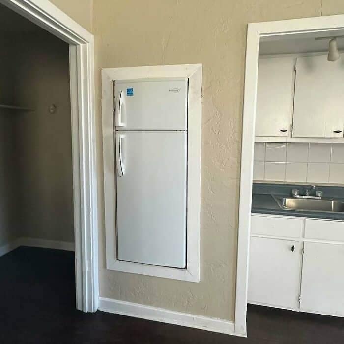 A standard white refrigerator framed with traditional thick door trim and mounted perfectly flush directly into a living room wall as a confusing piece of weird home decor.