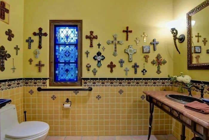 A yellow-tiled bathroom wall aggressively blanketed with dozens of mismatched religious crucifixes and decorative crosses, showcasing a surreal example of weird home decor.