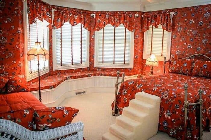 A bedroom heavily saturated in a matching red floral pattern across the wallpaper, window valances, curtains, and bedding sheets in a dizzying example of weird home decor.