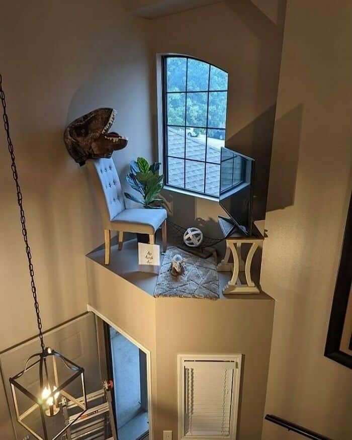 A narrow, completely un-railed interior architectural ledge high above a staircase drop-off, oddly furnished with a TV, a plant, and a chair holding a dinosaur mask in a surreal design fail.