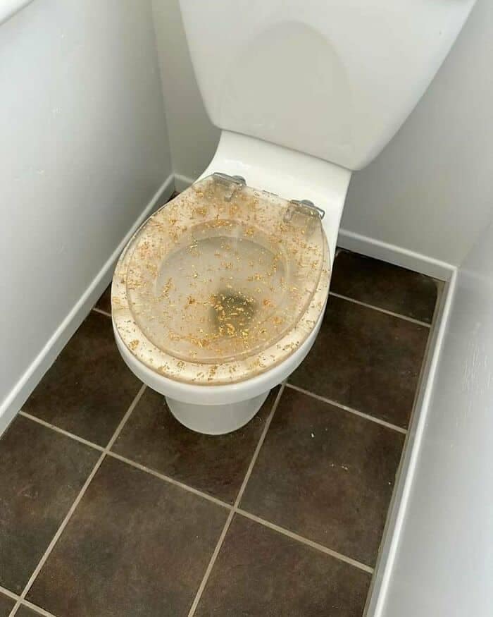 A close-up of a white toilet fitted with a clear resin seat embedded with gold leaf flakes that accidentally make the fixture look permanently stained and unsanitary in a catastrophic design fail.