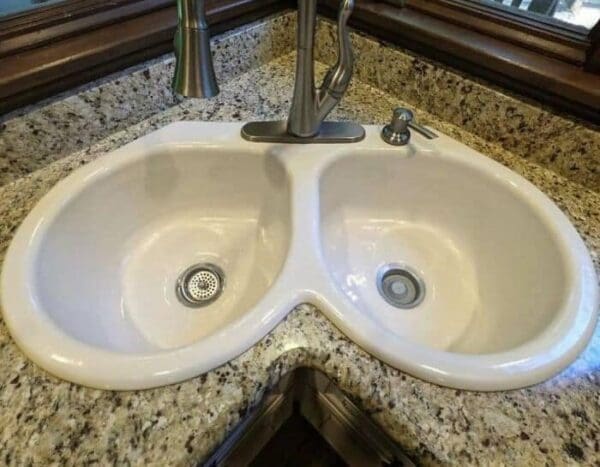 A white, double-basin corner kitchen sink uniquely sculpted into a rounded heart or butterfly-wing shape, installed tightly into a speckled granite countertop corner in a plumbing design fail.