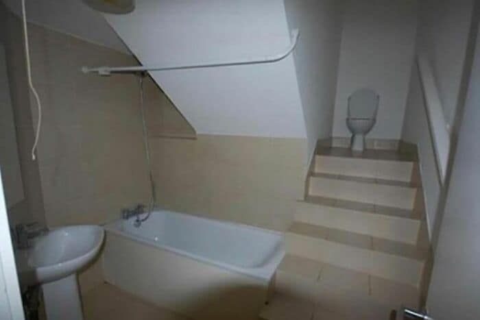 A bizarre bathroom featuring a tiled flight of stairs leading directly from the bathtub up to a lone toilet under a sloped ceiling, showcasing a classic design fail.