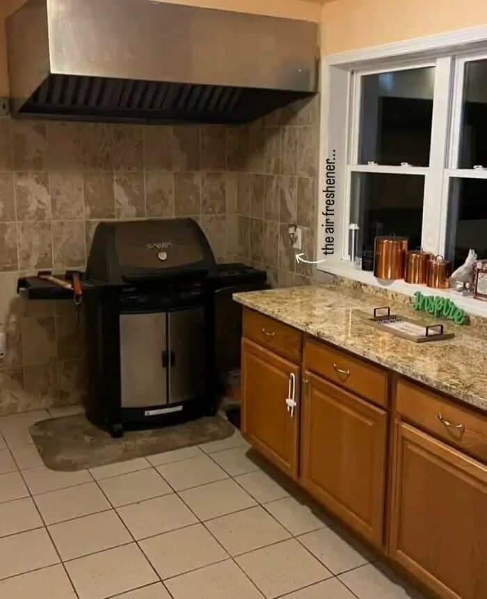 A residential kitchen featuring a full-sized outdoor stainless steel propane BBQ grill hooked up entirely indoors beneath a heavy sheet-metal commercial vent hood in an alarming example of weird home decor.