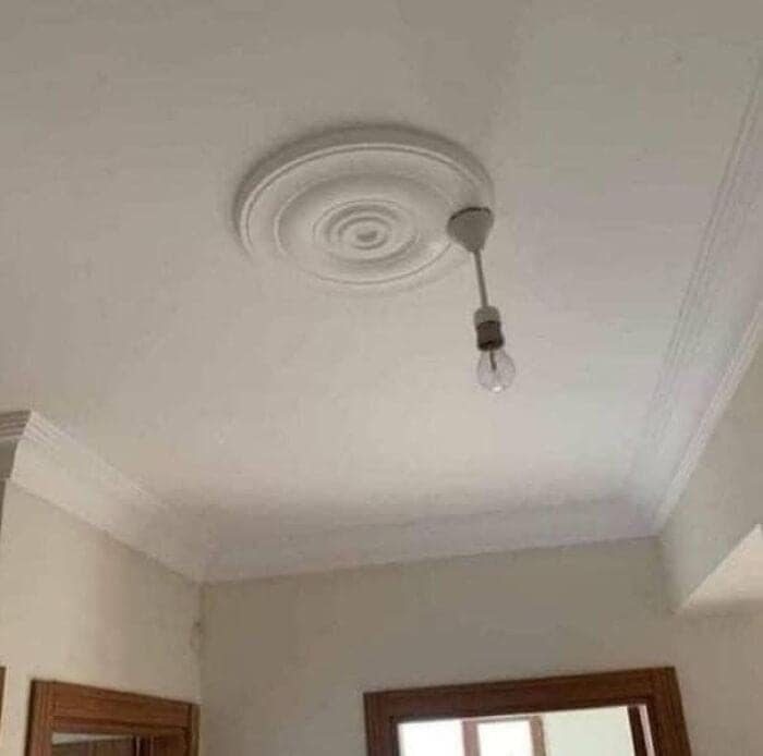A bare light bulb hanging completely off-center from the outer rim of a decorative plaster ceiling medallion instead of the middle hole, documenting a hilarious lighting design fail.
