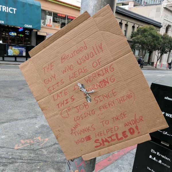 A city street photo serving as a community-focused wholesome meme, capturing a handwritten cardboard sign tied to a pole informing neighbors that the regular local unhoused man is absent because he finally landed a morning job.