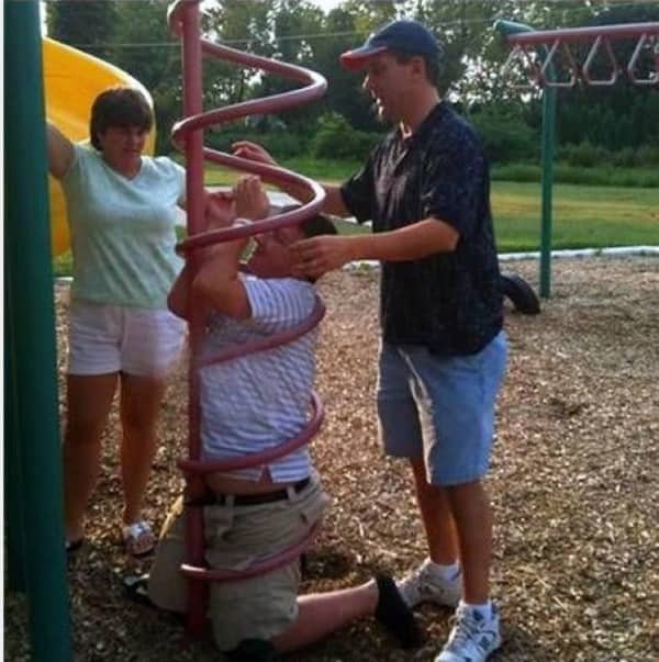 Grown man wedged inside a spiral playground ladder while two friends try to help him.