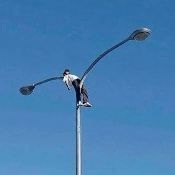 Person casually straddling the very top crotch of a double-sided street light pole.