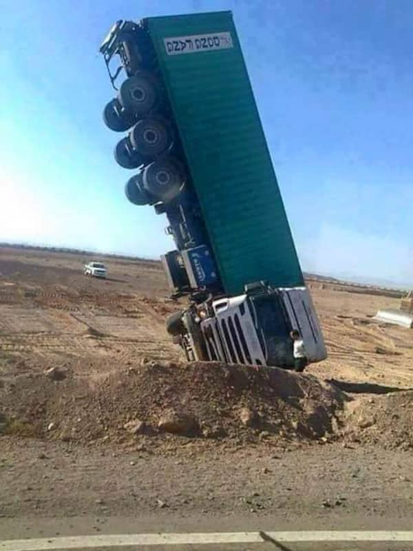 Massive green container truck standing entirely vertical on its front bumper in a dirt field.