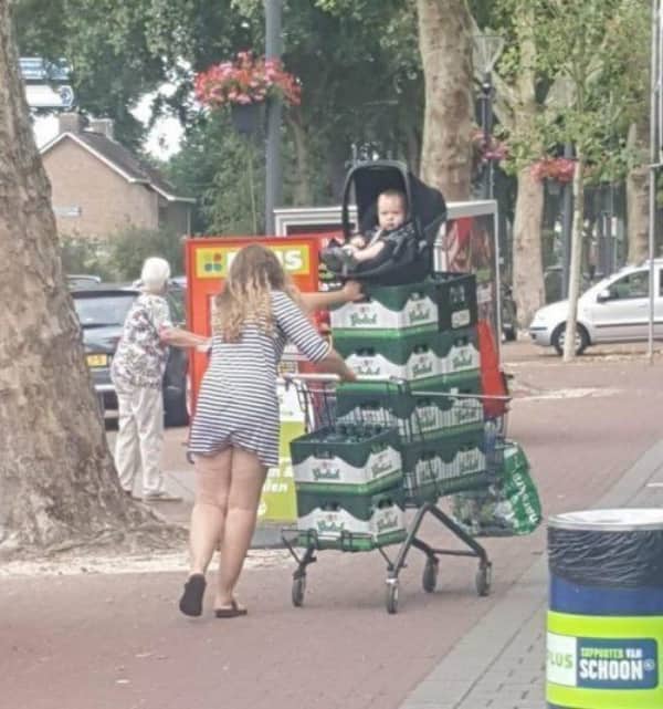 Woman pushing a shopping cart stacked with beer crates with a baby seat balanced on top.
